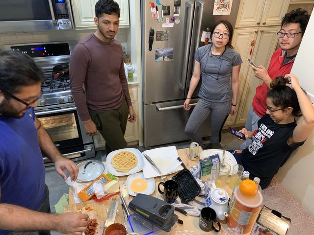 people in a kitchen surrounding a table of food